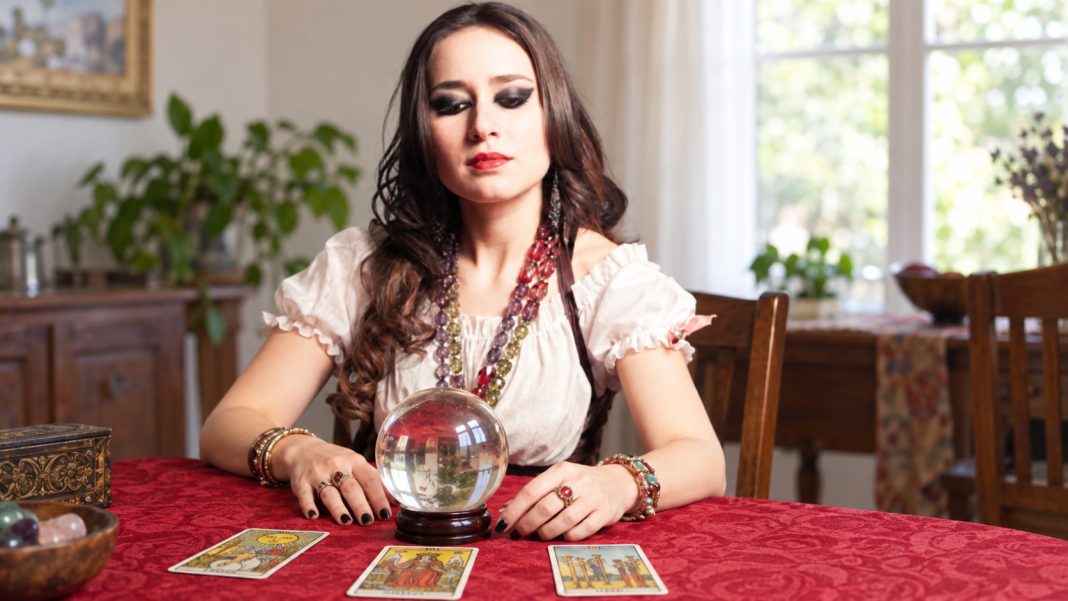 Illustrative reader in white blouse and layered jewelry at a home table with a clear sphere and tarot cards.