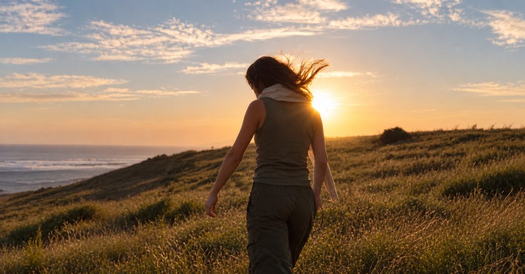 Woman seen from behind on a grassy coastal hillside at sunset.