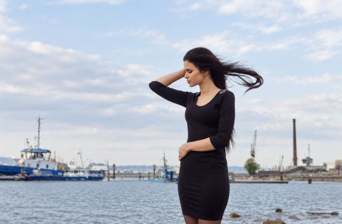 Past-life reflection: natural waterfront photograph Woman in a dark dress with windblown hair standing thoughtfully beside a harbor in daylight.