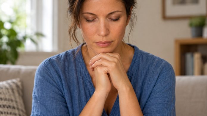 Psychic-arts introduction: realistic reflection portrait Woman in a blue shirt with eyes closed and hands together, seated in a softly lit home living room.