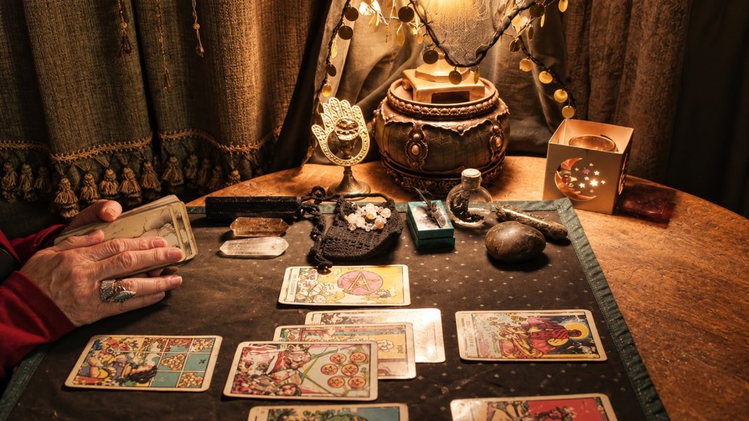 Reader's ringed hands holding a card beside a tarot spread on a wooden table.