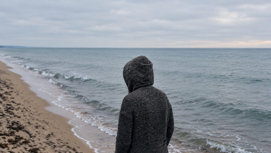 Hooded person seen from behind looking toward the sea.