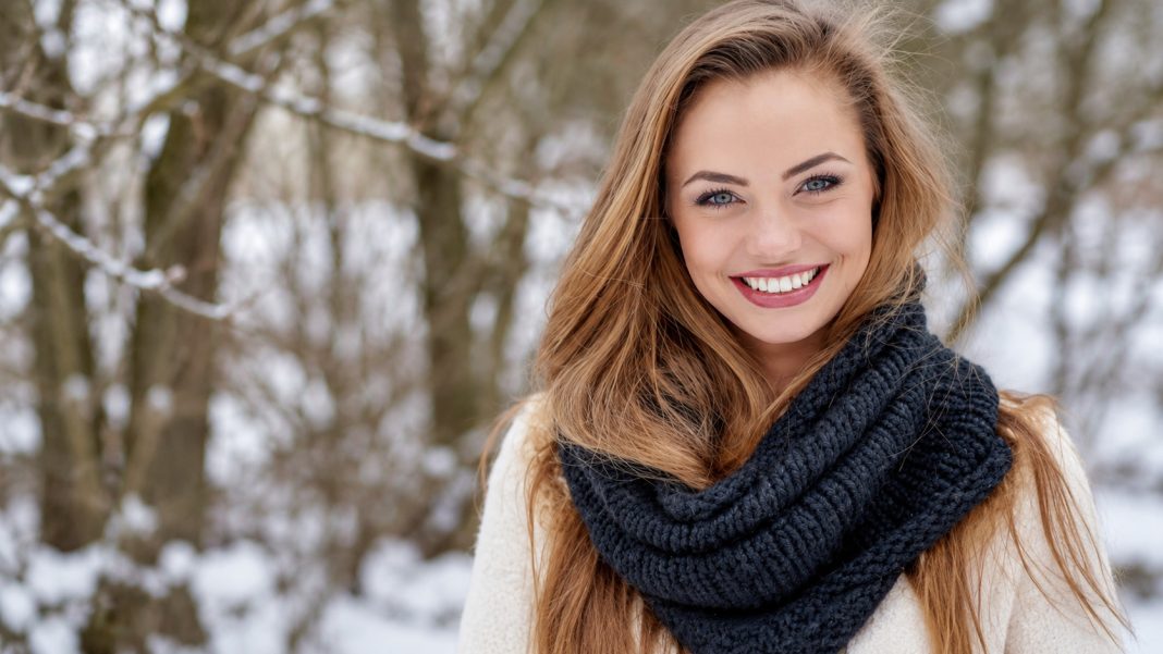 Smiling woman in a dark knitted scarf and pale outer clothing outdoors among trees.